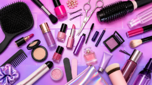 Flat‑lay of makeup products, brushes, hair tools, and beauty accessories arranged on a pink and purple background